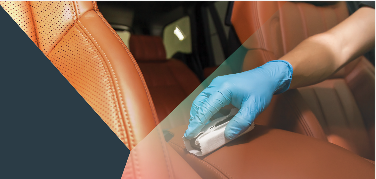 A close-up of a person wearing a blue nitrile glove wiping down an orange leather car seat with a small white cleaning cloth.