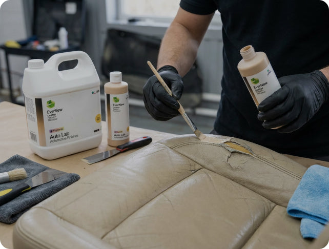 A person wearing black gloves applies a liquid restoration product from a bottle with a brush onto a damaged beige leather seat, with Auto Lab bottles and tools visible on a workbench.