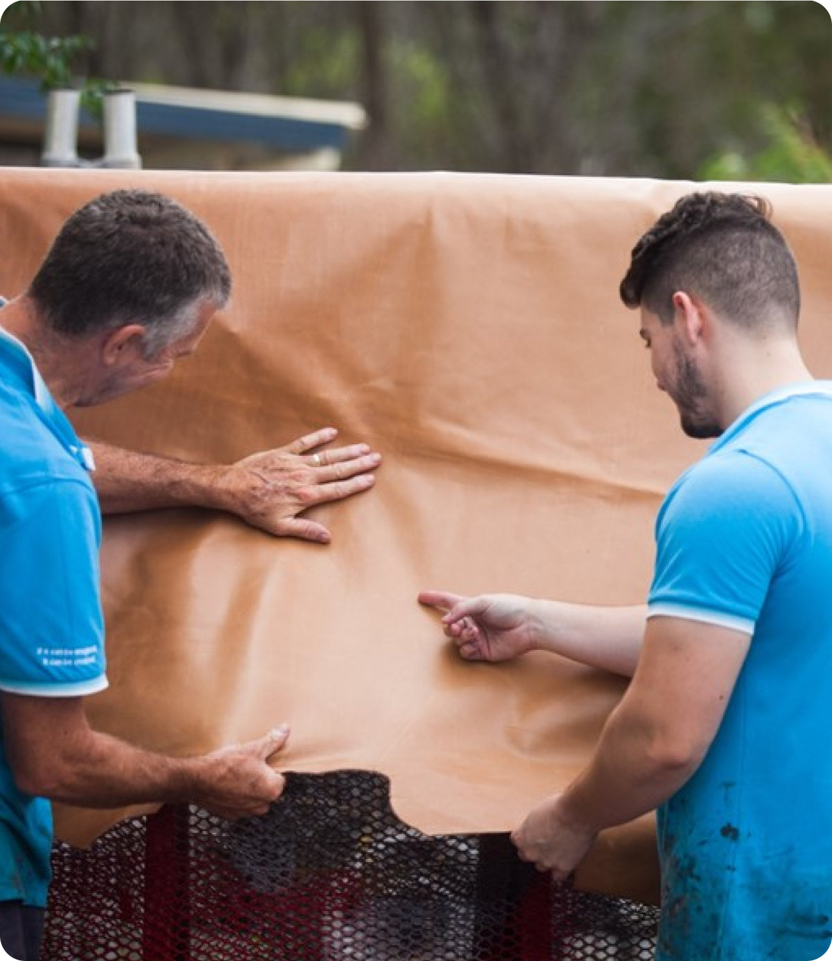 Darryl and a trainee handling a large tan or light brown sheet or mat outdoors, with trees and a structure blurred in the background.