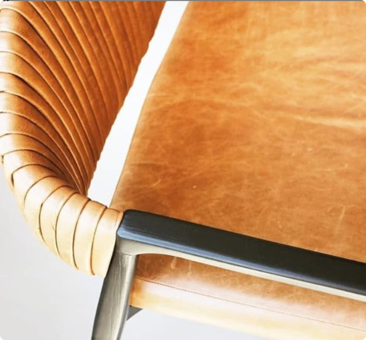 Close-up of a modern chair with a curved, pleated tan leather backrest, a matching tan leather seat, and a black metal armrest.