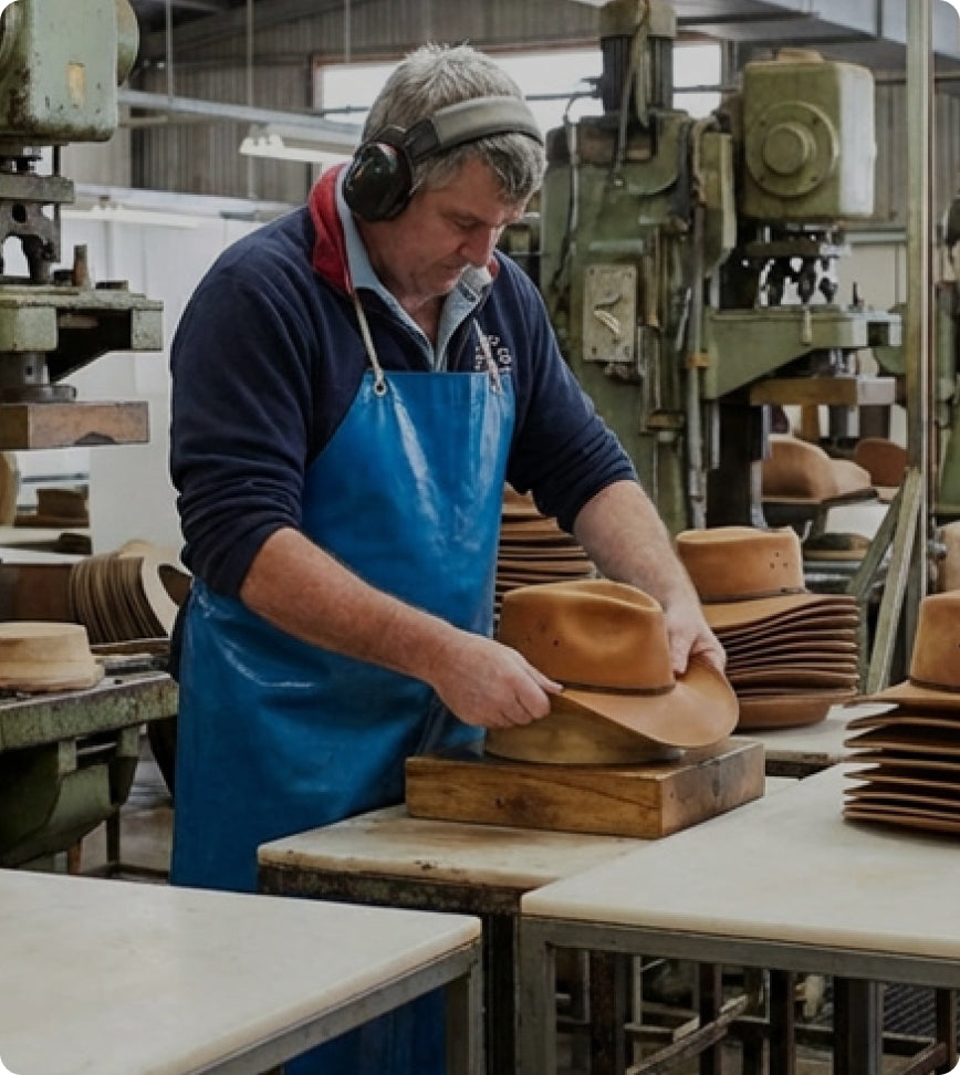 A man wearing over-ear headphones and a blue apron works at a table in a hat manufacturing workshop, shaping a tan cowboy hat on a wooden press. Stacks of finished leather hats are visible in the background.