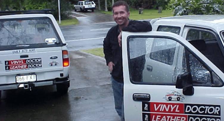 Darryl Binch smiling and leaning on the open door of a white vehicle with 'VINYL DOCTOR LEATHER DOCTOR' decals, standing outdoors on a wet road next to another white vehicle.