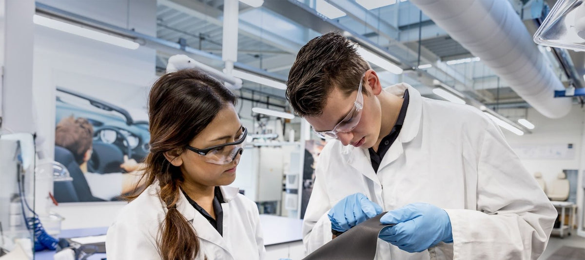 Two scientists in white lab coats and safety glasses examining a tablet in a modern laboratory setting.
