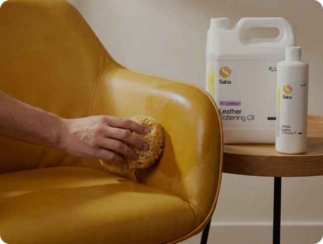 A person's hand uses a sponge to apply Saba Leather Softening Oil from a white bottle and jug onto a mustard yellow leather chair, with a small wooden side table holding the products in the background.